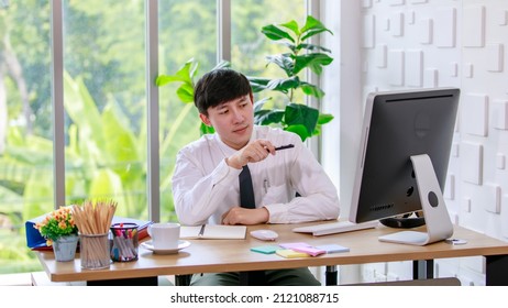 Portrait Studio Shot Of Asian Professional Successful Male Businessman Employee In Formal Shirt With Necktie Sitting Look At Camera At Working Desk With Computer Monitor Keyboard Mouse And Stationery.