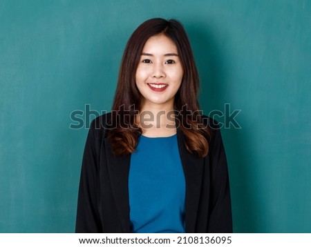 Portrait studio shot of Asian professional long hair female teacher or college student in formal suit standing smiling look at camera hold hands in jacket pockets on green chalkboard background.