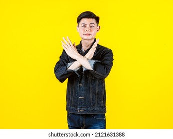 Portrait studio shot of Asian unsatisfied young male model in casual jeans jacket look at camera showing crossed hands symbol sign say no reject refuse deny on yellow background with copy space.