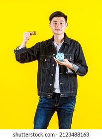 Portrait Studio Shot Of Asian Young Rich Wealthy Male Hipster Model Wearing Casual Street Denim Jeans Jacket Look At Camera Holding Showing Golden Coin And Piggy Bank Saving On Yellow Background.