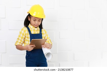 Portrait studio shot Asian young little elementary schoolgirl future dream job career as engineer and architect wears yellow safety hardhat helmet standing look at camera holding tablet and blueprint.
