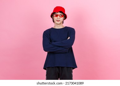 Portrait Studio Shot Asian Young Urban Teen Fashion Male Model In Street Style Outfit Long Sleeve Shirt Red Bucket Hat Funny Glasses Standing Crossed Arms Smiling Look At Camera On Pink Background.