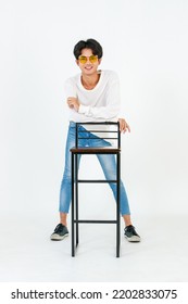 Portrait Studio Shot Of Asian Young LGBT Gay Happy Bisexual Homosexual Male Fashionable Model In Casual Outfit Fashion Sunglasses Standing Leaning At Tall Chair Smiling Posing On White Background.