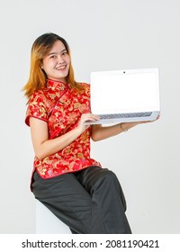 Portrait Studio Shot Millennial Asian Female Model In Red Chinese Traditional Cheongsam Qipao Shirt Sitting Holding Showing Presenting Blank Screen Laptop Computer Product On White Background.