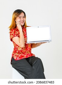 Portrait Studio Shot Millennial Asian Female Model In Red Chinese Traditional Cheongsam Qipao Shirt Sitting Holding Showing Presenting Blank Screen Laptop Computer Product On White Background.