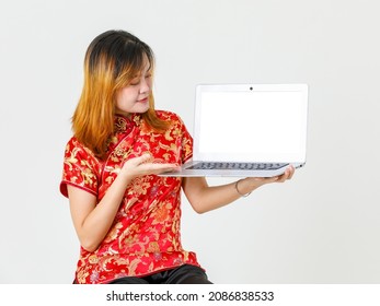 Portrait Studio Shot Millennial Asian Female Model In Red Chinese Traditional Cheongsam Qipao Shirt Sitting Holding Showing Presenting Blank Screen Laptop Computer Product On White Background.