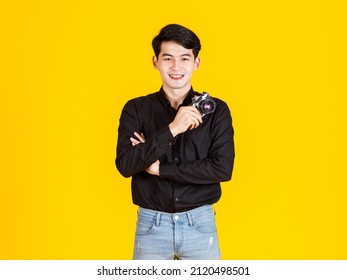 Portrait studio shot of millennial Asian young professional male fashion photographer journalist in casual outfit standing smiling holding aiming digital DSLR mirrorless camera on yellow background. - Powered by Shutterstock