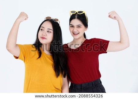 Portrait studio shot of two young lovely Asian female friend sister model in casual t shirt outfit and fashion sunglasses standing smiling showing strong biceps muscle muscular on white background.