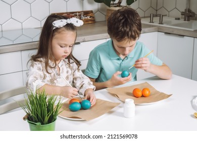 Portrait of studiously two little children, boy and girl, painting brush multi coloring Easter hens eggs on paper plate table, plant in kitchen. Making white art patterns. Traditional holiday event.