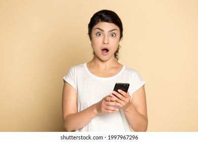 Portrait of stunned Indian woman isolated on yellow studio background use cellphone shocked by news online. Amazed young mixed race female surprised by unexpected sale deal or discount on smartphone.