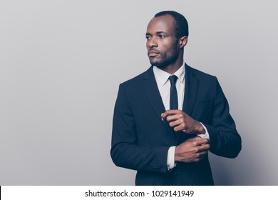 Portrait of stunning, trendy, attractive, dreamy, perfect man in black suit with tie fasten button on sleeve cuffs of white shirt, looking to the side, isolated on grey background