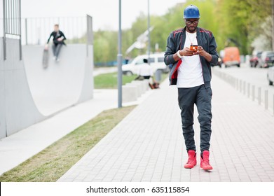 Portrait of stylish african american man on sportswear, cap and glasses looking on his phone. Black men model walk at skate park.
