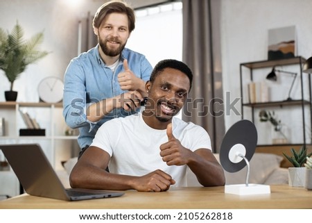 Portrait of stylish African man looking into the camera and showing thumb up while his Caucasian friend shaving his neck with an electric razor to create a stylish hairstyle. Multiracial gay couple