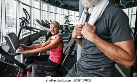 Portrait Of Stylish Attractive Aged Woman In Pink Top Working Out On Gym Cycle, Looking At Camera Next To Unrecognised Man In Gray T-shirt. Selective Focus On Woman. Gym With Large Windows.