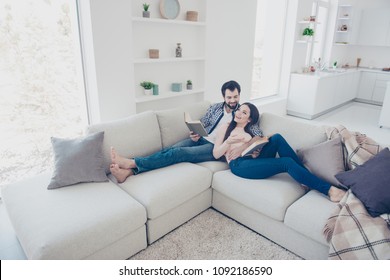 Portrait of stylish attractive couple enjoying reading novel, poem, poetry, lying on sofa having books in hands, woman telling showing comic plot from her detective story