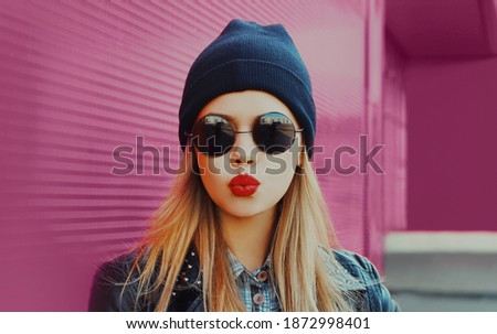 Portrait of stylish beautiful blonde woman blowing red lips sending sweet air kiss wearing a black rock style over a pink background