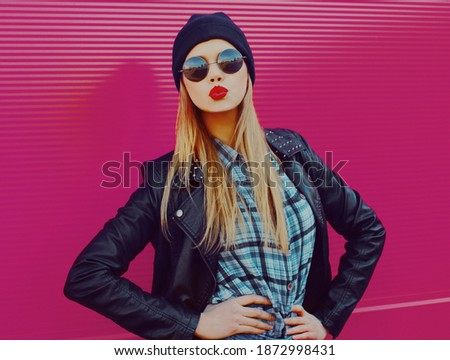 Portrait of stylish beautiful blonde woman blowing red lips sending sweet air kiss wearing a black rock style over a pink background