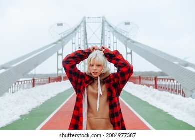 Portrait of a stylish blonde girl in a red shirt on the background of the bridge with snow in the winter, posing for the camera