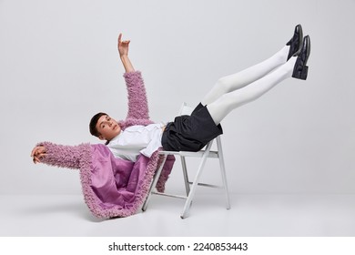 Portrait of stylish boy in pink furry coat, white tights, black shorts lying on chair, posing isolated over grey background. Freedom. Concept of modern fashion, art photography, style, uniqueness, ad