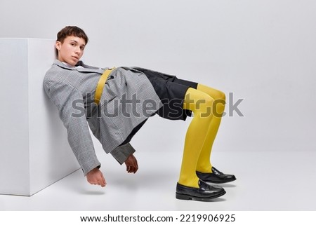 Portrait of stylish boy posing in jacket, blazer and yellow tights isolated over grey background. Leaning on cube. Concept of modern fashion, art photography, style, queer, uniqueness, ad
