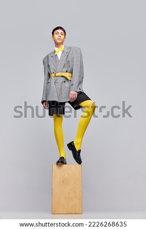 Portrait of stylish boy posing in jacket, blazer and yellow tights and bow isolated over grey background. Statue style. Concept of modern fashion, art photography, style, queer, uniqueness, ad