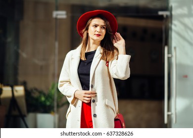 Portrait Of A Stylish Brunette Business Woman With A Red Bag In Hands Wearing In White Coat Red Hat, Red Pants And Shoes Standing At The Entrance Of Hotel Or Boutique.