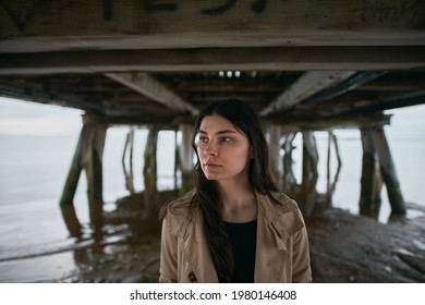 Portrait Of Stylish Brunette Under The Wooden Pier. Attractive And Trendy Girl With Long Brown Hair By The Seaside Stand In Tan Color Trench Coat.