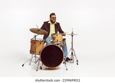 Portrait of stylish brutal man in jacket and sunglasses playing drums isolated over white background. Concept of live music, performance, retro style, creativity, artistic lifestyle