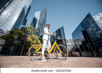 Portrait of a stylish business woman in white suit standing with bicycle at the financial district with modern buildings on the background