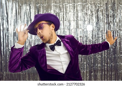 Portrait of stylish and cool young showman, cheerful partygoer or glamorous visitor of stylized party. Dark-skinned young man in purple corduroy suit dances and has fun on silver shiny background.