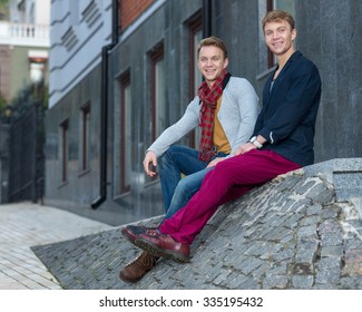 Portrait of stylish fashionable twin brothers sitting on the stairs with cheerful emotions. Twins outdoor fashion style concept.