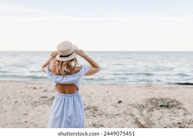 Portrait of stylish female on sand. Beautiful girl standing on beach ocean and enjoying sunny summer happy day on vacation. Back view. Young attractive blond woman in straw hat joyfully walks near sea - Powered by Shutterstock