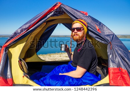 portrait of stylish ginger red-haired beard tourist man in a colorful hat made of yak wool from Nepal holding a mug of tea coffee in tent wrapped sleeping bag the background lake