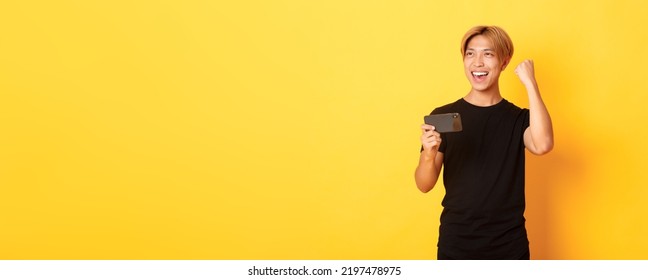 Portrait Of Stylish Handsome Asian Guy Playing Smartphone Game, Rejoicing From Winning, Standing Yellow Background