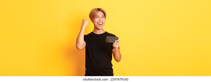 Portrait Of Stylish Handsome Asian Guy Playing Smartphone Game, Rejoicing From Winning, Standing Yellow Background