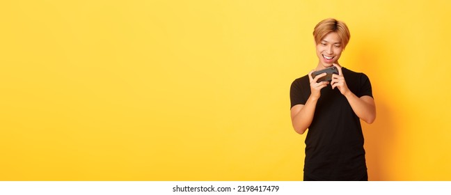 Portrait Of Stylish Handsome Asian Guy Playing Smartphone Game, Looking Amused At Mobile Screen, Standing Yellow Background