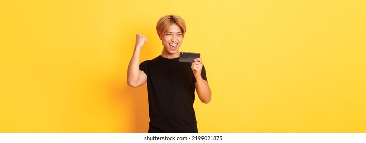 Portrait Of Stylish Handsome Asian Guy Playing Smartphone Game, Looking Amused At Mobile Screen And Rejoicing From Winning, Standing Yellow Background