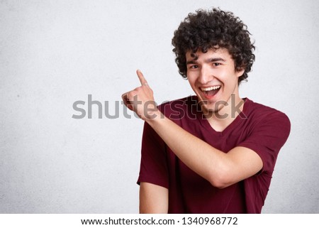 Portrait of stylish handsome young guy dresses casual outfit, with dark curly hair isolated on white background. Laughing teenager being in good mood points up. People, youth and lifestyle concept.