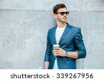 Portrait of stylish handsome young man with bristle standing outdoors and leaning on wall. Man wearing jacket, sunglasses, shirt and holding cup of coffee