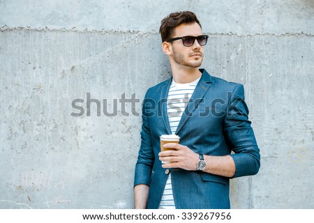 Portrait of stylish handsome young man with bristle standing outdoors and leaning on wall. Man wearing jacket, sunglasses, shirt and holding cup of coffee