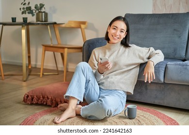 Portrait of stylish korean woman sits on floor with smartphone, using mobile phone, smiling pleased, concept of staying at home and relaxation. - Powered by Shutterstock