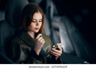  portrait of a stylish, luxurious woman in a leather coat sitting in a black car at night in the passenger seat, thoughtfully looking at her phone during the trip, holding her hand near her face - Powered by Shutterstock