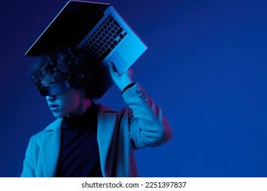 Portrait of a stylish male hacker with a laptop and futuristic glasses in blue light, cyber security, technology and education, neon color, freelancer online work, trend color