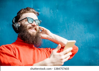 Portrait of a stylish man dressed in red sweater enjoying music with headphones and smart phone on the blue background