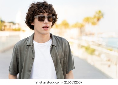 Portrait Of Stylish Man Wearing Sunglasses Looking Away Outdoor. Curly Guy Feeling Happy And Confident At Sunset. Closeup Face Of Boy Smiling With Street In Background 