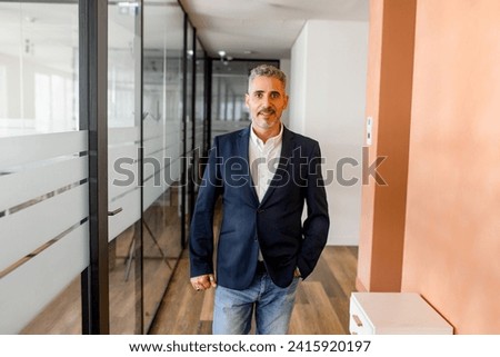 Portrait of a stylish mature businessman standing in the modern office and posing against the glass partition, male employee, manager looking at the camera