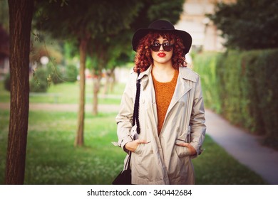 Portrait Of Stylish Modern Young Curly Redhead Woman In Trench Coat, Sunglasses And Fedora Hat Standing In Park In Autumn. Horizontal, Retouched, Color Filter Applied.