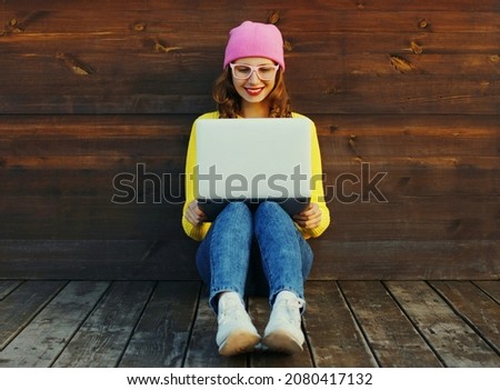 Portrait of stylish modern young woman working with laptop wearing a colorful clothes sitting in the city park on wooden wall background