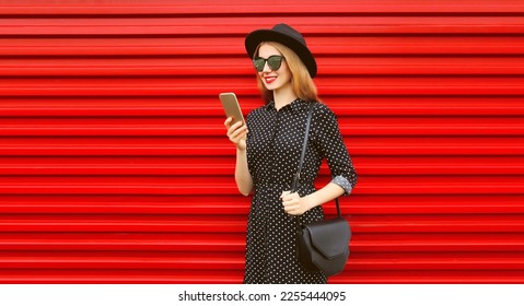 Portrait of stylish smiling woman with smartphone wearing black round hat on red background - Shutterstock ID 2255444095