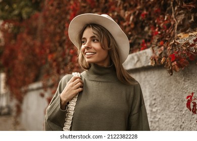 Portrait of stylish smiling young woman with blond hair walking in park, wearing warm sweater. Girl looks back towards her acquaintances. Autumn trendy fashion, street style with hat.